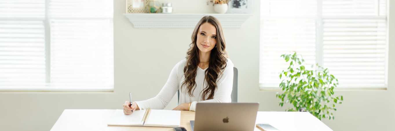 Taylor Ann, someone with NMOSD, smiling at her desk with a laptop and notebook.